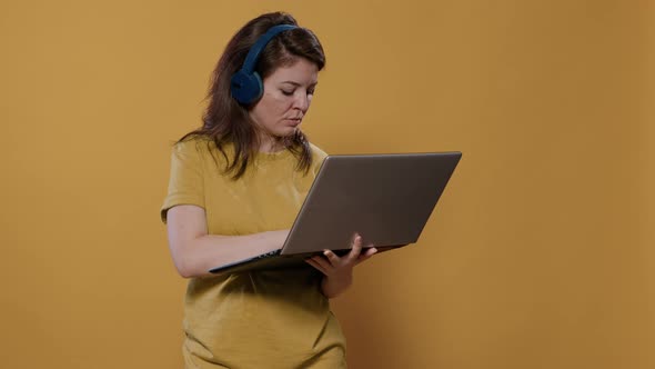 Casual Woman Wearing Wireless Headphones Holding Laptop and Typing on Keyboard Looking at Screen