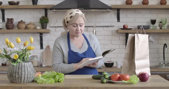 Positive Caucasian Senior Woman Looking for New Salad Recipe Standing in Kitchen. Portrait of