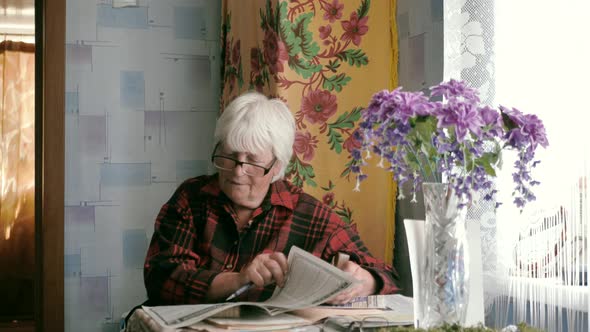 Portrait of an Old Woman in Glasses Reading a Newspaper While Sitting at a Table at Home