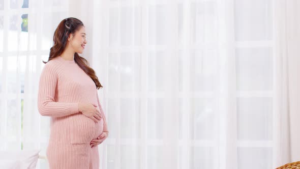 Happy Pregnant Woman standing in front of windows and stroking her big belly with love at cozy home