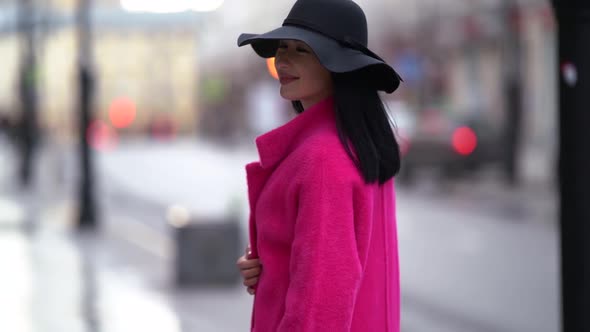 Portrait of a Pretty Black-haired Woman in a Pink Coat and Black Hat Walking on a Blurry Background