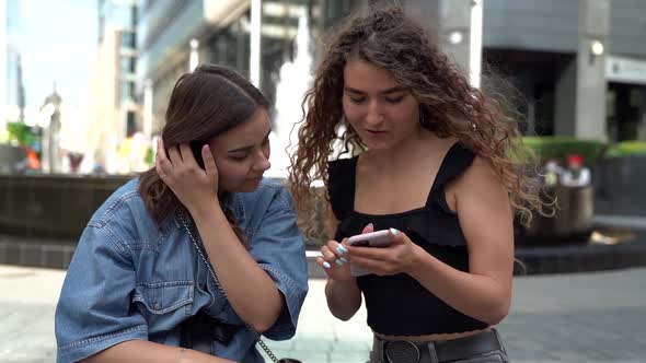 Two Young Female Friends Are Viewing Pictures in Smartphone, Sitting in City at Summer Day