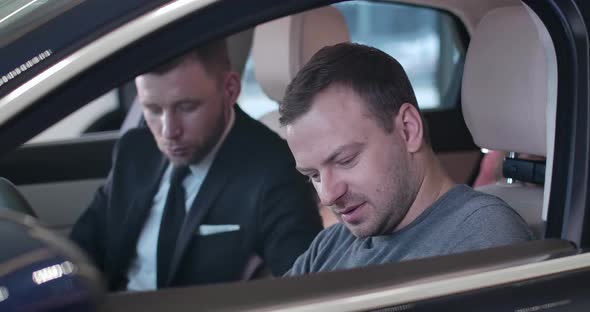 Close-up of Caucasian Young Man Sitting in Car Salon with Blurred Dealer at the Background. Trader