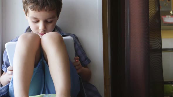 Cute Little Boy Uses A White Tablet Pc On A Windowsill At Home