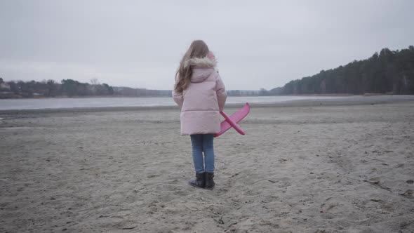 Back View of Caucasian Schoolgirl in Pink Clothes Standing in Wind on the Riverbank and Holding Toy