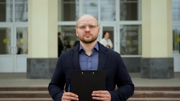 Young Man Stands Against the Background of the University with a Folder in Hands