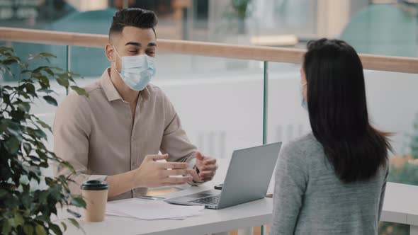 Young Hispanic Bank Worker in Medical Face Mask Telling Unrecognizable Client Woman Benefits Using