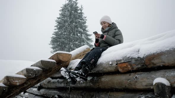 Young Woman Use Mobile Phone Under a Snowfall in the Mountains at a Resort Sitting on the Snow