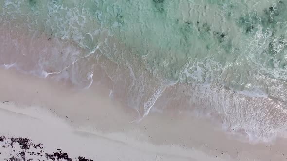Aerial view looking down of clear sea waves crashing onto a white sand beach while drone moves towar