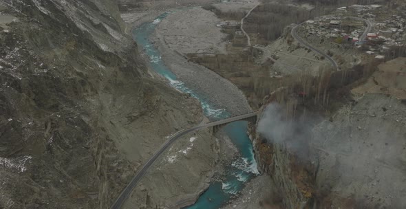 Aerial Over Turquoise Colour River In Hunza Valley With Smoke Seen Rising Near Bridge. Dolly Zoom Ti