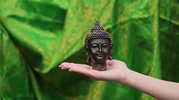 Young woman hand with long nails holding a Buddha head statue in front of green background.