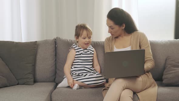 Mother and Her Young Daughter Spend Time Together Sitting on a Couch with a Laptop