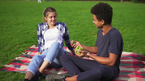 Young Lovely Couple Eating Fruit Sitting on the Grass in Park on Summer Day