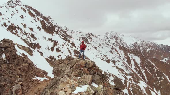 A Male Traveller with a Backpack Stands on Top of a Cliff Against the Backdrop of Snowcapped