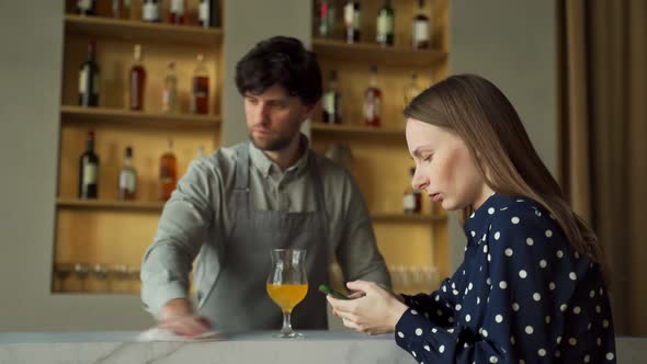 Woman Using a Smartphone at the Bar Against the Background of a Bartender Wiping the Table