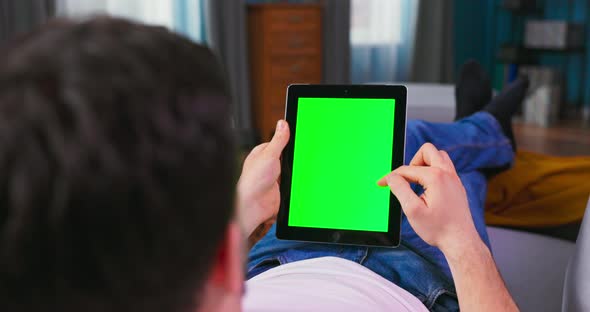 Young Man at Home Resting on a Couch Using with Green Mockup Screen Tablet Computer