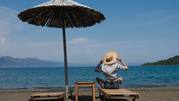 Girl in Shirt and Hat Sitting on the Beach on a Sunbed Under an Umbrella