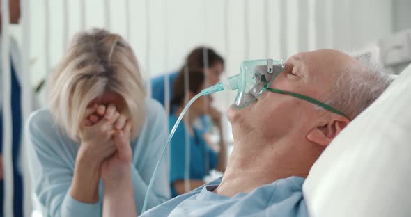 Crying Woman Holding Hand of Aged Husband Patient While Sitting By Bed at Hospital