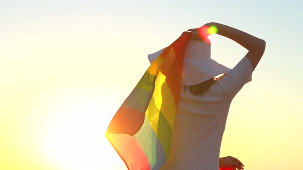 Back View of Woman in White Dress and Hat Holding Gay Pride Flag at Sunset in Beach