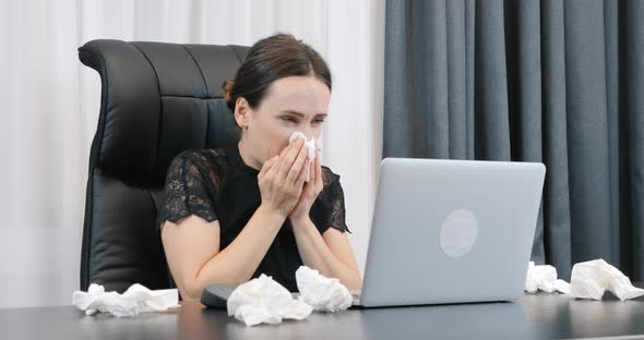 Sick woman with runny nose working in office with dirty paper napkins lying around