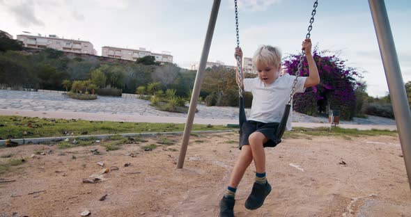 Cute baby blond boy swinging on chain swing in playground in flowering park