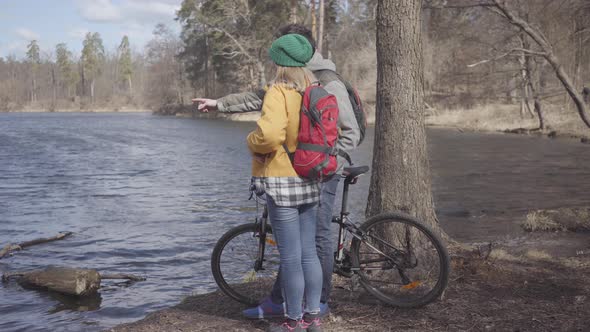 Young Couple of Tourists Standing on the Riverbank with Backpacks on Their Backs. The Man Holds the