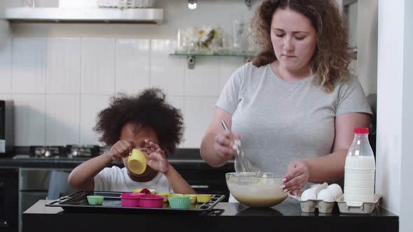 Little Black Girl Looking in the Moulds While Her Mom Stirs the Dough with a Whisk