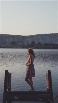 Young woman in dress looking at camera while standing on wooden platform in calm lake in mountains