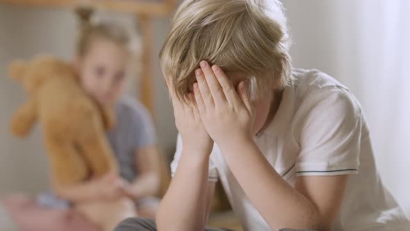 Portrait of Playful Caucasian Boy Closing Face with Hands and Grimacing with Blurred Girl Sitting at