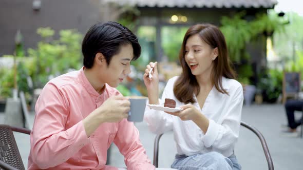 Young asian woman feeding happy boyfriend with tasty chocolate cake sitting at table with coffee 