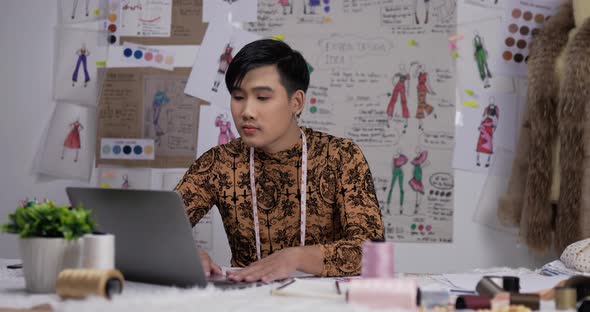 Asian clothing designer working on laptop in his office, creating a new clothes collection