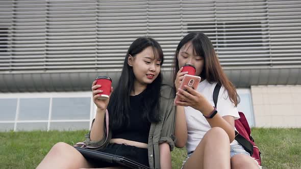 Two Pleasant Cheery Asian Brunettes which Sitting on Grass and Using their Phones