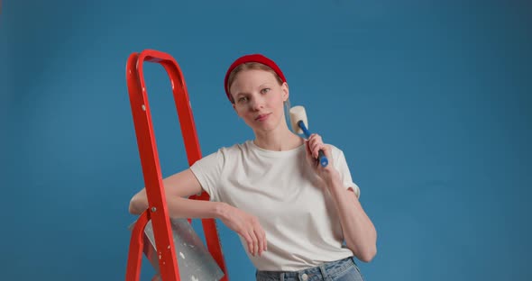 Young Funny Woman in Red Cap Stands with Stepladder Holding Hammer in Hand on Blue Background