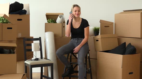 A Happy Moving Woman Sits on a Chair in an Empty Apartment and Talks on a Smartphone with a Smile