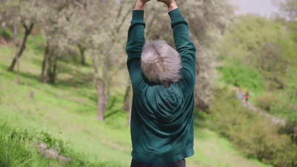 Back View of Satisfied Senior Greyhaired Caucasian Woman Raising Hands Up Stretching in Sunny Park