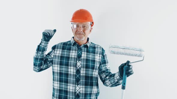 A Cheerful Grayhaired Man in a Hard Hat and Protective Glasses Holds a Roller with Paint in His Hand