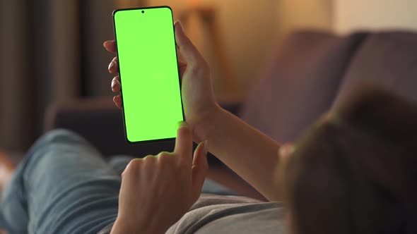 Woman at Home Lying on a Sofa and Using Smartphone with Green Mock-up Screen in Vertical Mode. Girl