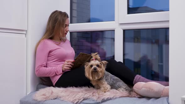 Young Woman Sits on Windowsill Near Large Windowenjoys Reading an Interesting Book