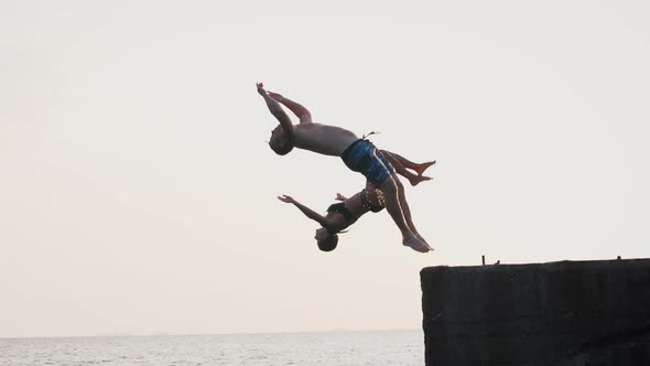 Young Woman and Man Synchronously Doing Trick Jump From a Pier Into the Sea During Beautiful Sunrise