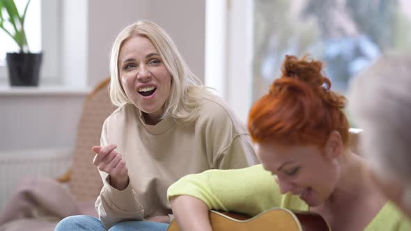 Portrait of Joyful Blond Beautiful Adult Woman Singing As Friend Playing Guitar Sitting on Couch at