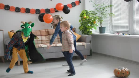 Halloween Party Boy in Dragon Costume Runs with Friends in a Room Decorated with Pumpkins Garlands