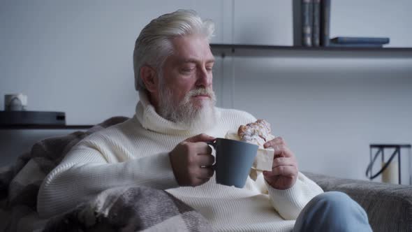 Elderly Man with a Gray Beard Is Sitting on a Chair Drinking Coffee and Eating a Croissant