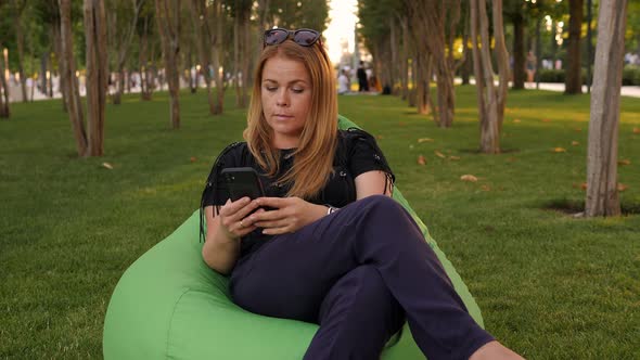 A Young Woman Sits in an Urban Modern Crowded Park in a Soft Chair with a Phone