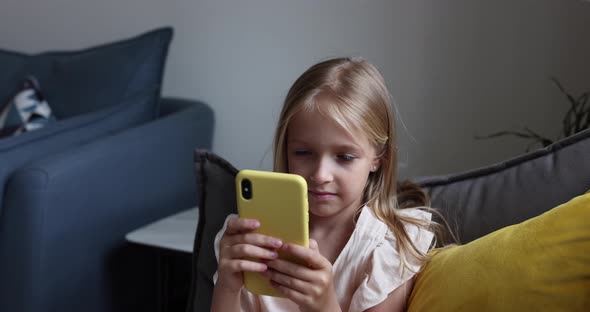 Happy Little Caucasian Girl with Blonde Hair Seven Years Old Using Smartphone on Sofa At Home