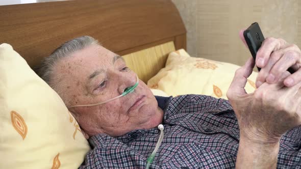 Old Man Lies in Bed with an Oxygen Tube and Looks at Phone Screen