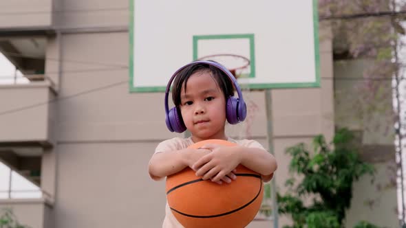 Cute little girl wearing headphones poses with basketball at outdoor basketball court. alt