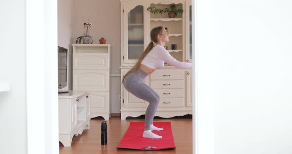 Side view of an attractive girl squatting on the red mat doing sports exercises indoors at home 