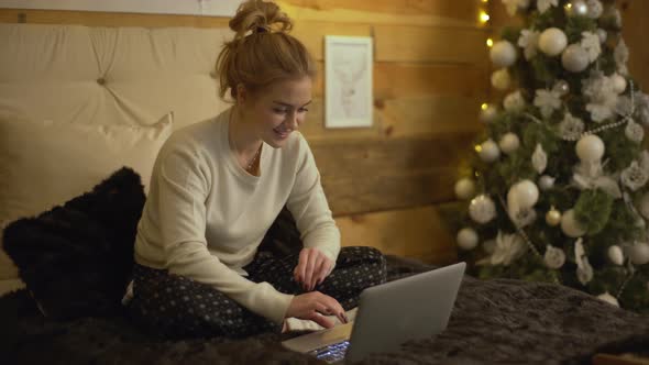 Portrait of Pleased Blond Woman Sitting on Bed with Legs Crossed in Cozy Room with Christmas Tree