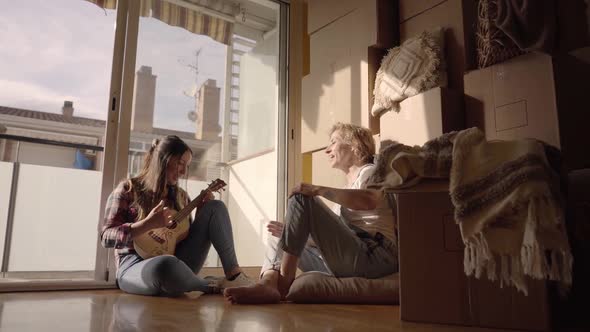 Happy Women Landlords Tenants Sit on the Floor Play Ukulele and Sing on Moving Day in New House