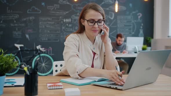 Blond Girl Working with Laptop in Office Typing and Speaking on Mobile Phone